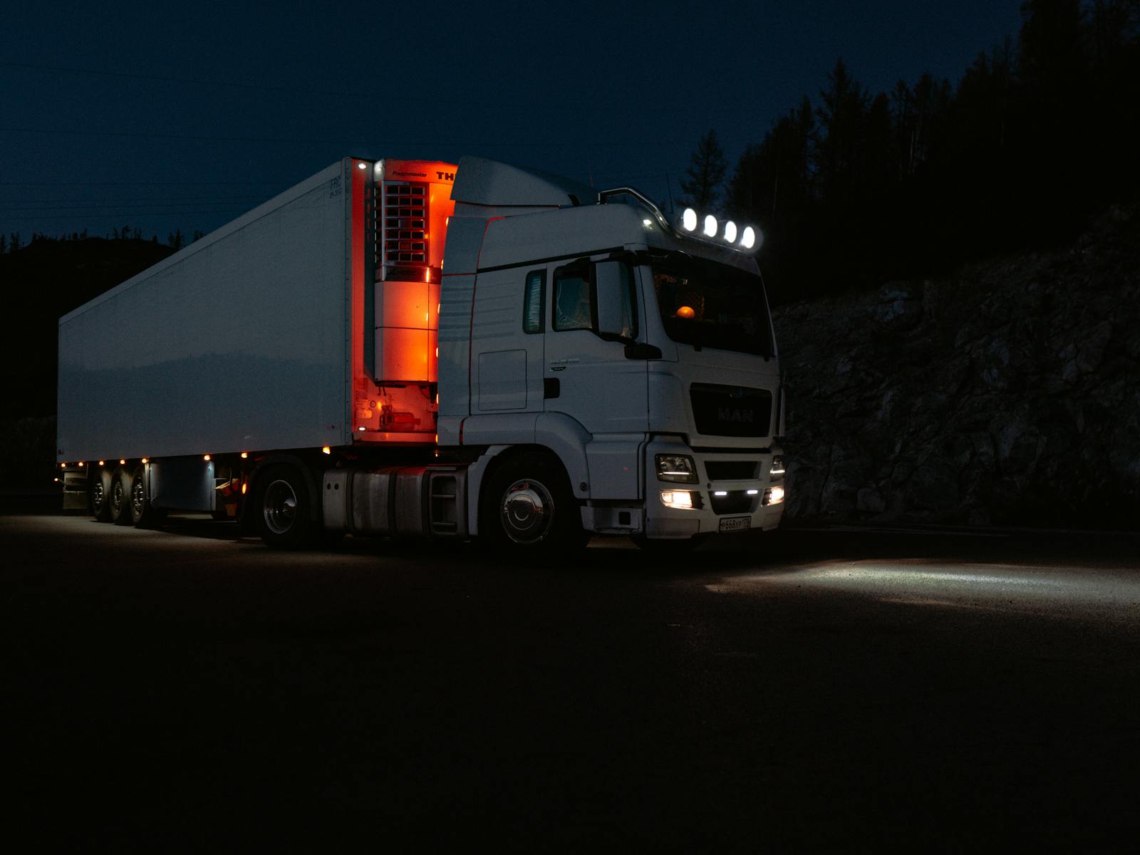 trucking, white truck lit by headlights and interior lights on a dark road in mountainous terrain.